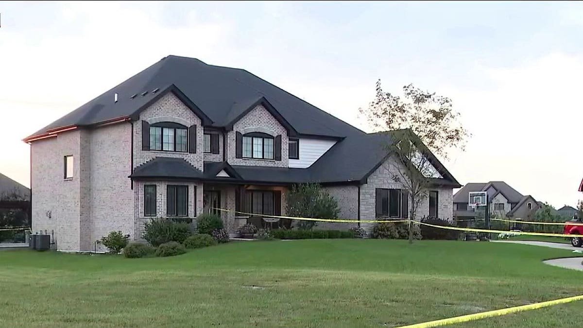 A large two-story brick home sits behind yellow police tape in a residential neighborhood.