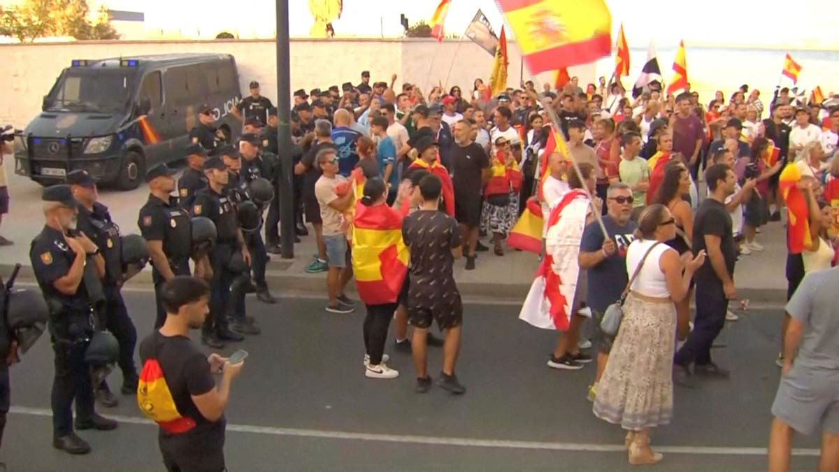Law enforcement personnel watch while protesters hold Spanish flags, as Ceuta residents destroy migrant camp by the beach