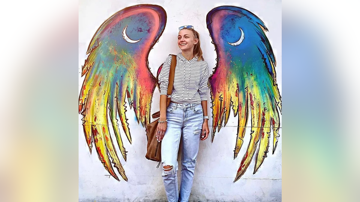 A portrait of Gabby Petito wearing a striped shirt and bluejeans standing in front of painted angel wings