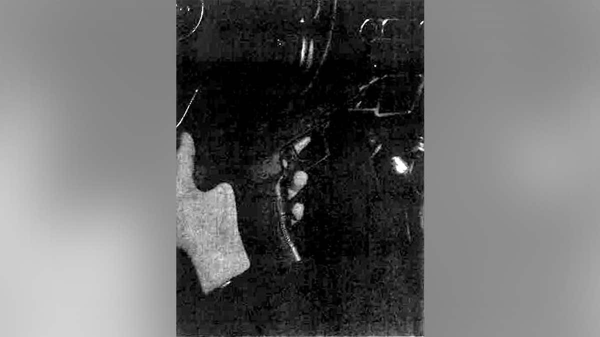 Grainy black-and-white still showing a person in a light-colored shirt holding an object near a residential property.