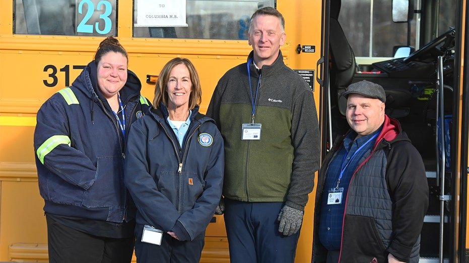 Kurtis Kotes stands with school transportation staff in front of a yellow school bus.